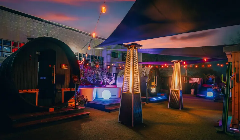 Outdoor courtyard at Bathe at dusk with a barrel sauna, patio heaters, string lights and a shade sail