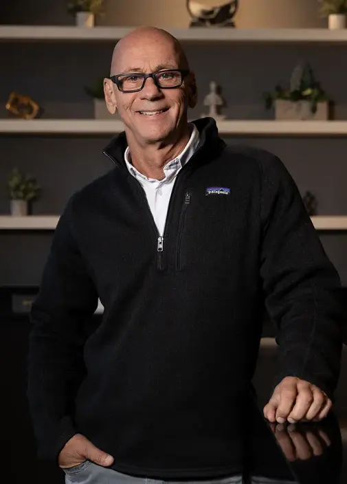 Portrait of Dr. Gladden in glasses and a black fleece, standing in front of a shelved wall