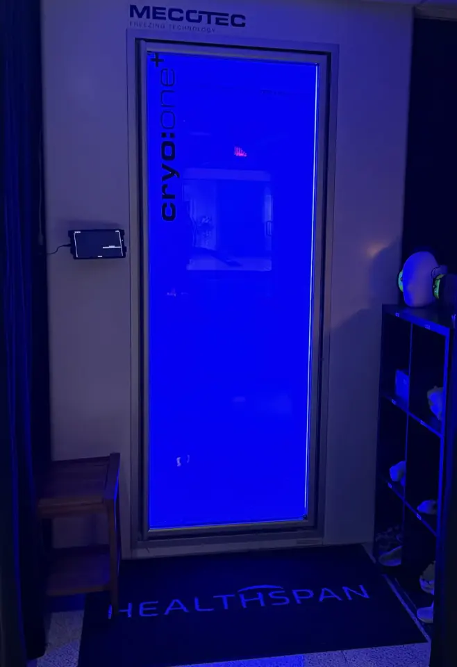 Whole-body cryotherapy chamber glowing blue above a Healthspan floor mat