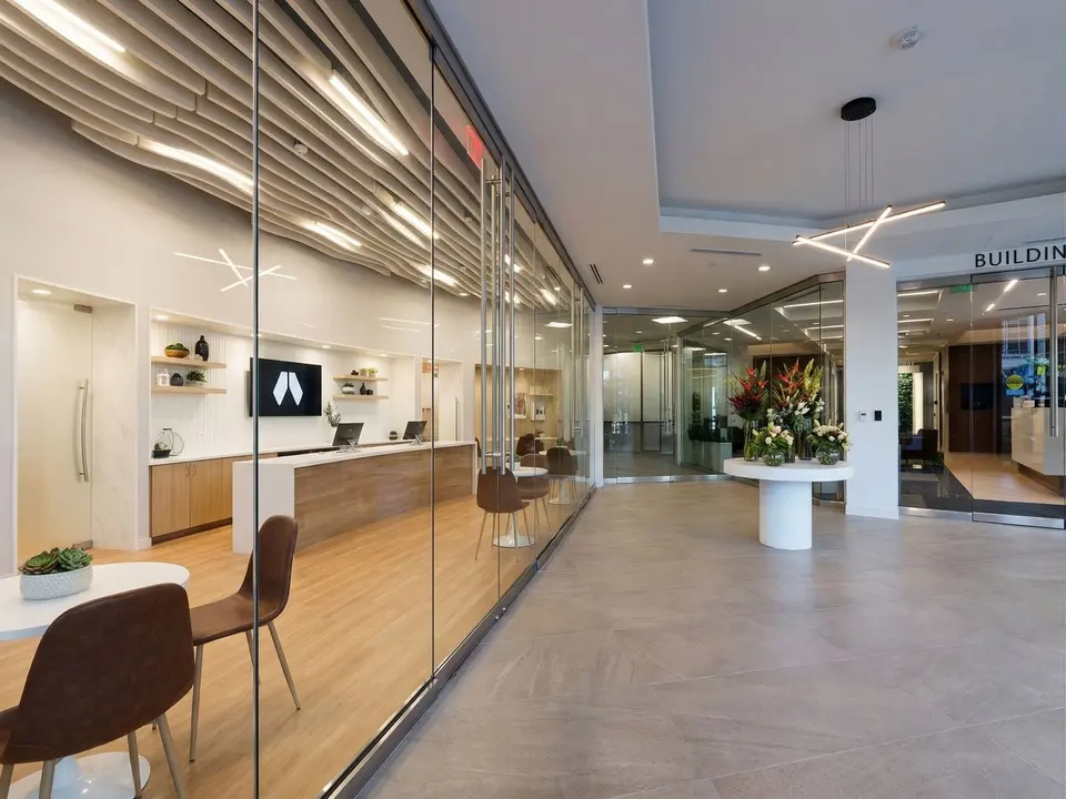 Glass-walled clinic interior at Humanaut Health Dallas Preston Center with wood-slat ceiling, reception counter and a flower arrangement in the corridor