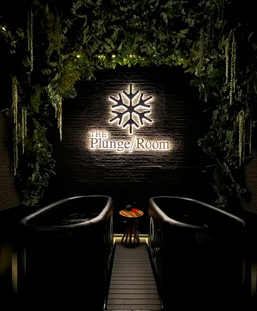 Two cold plunge tubs beneath a backlit Plunge Room sign framed by hanging greenery in the Hoboken suite