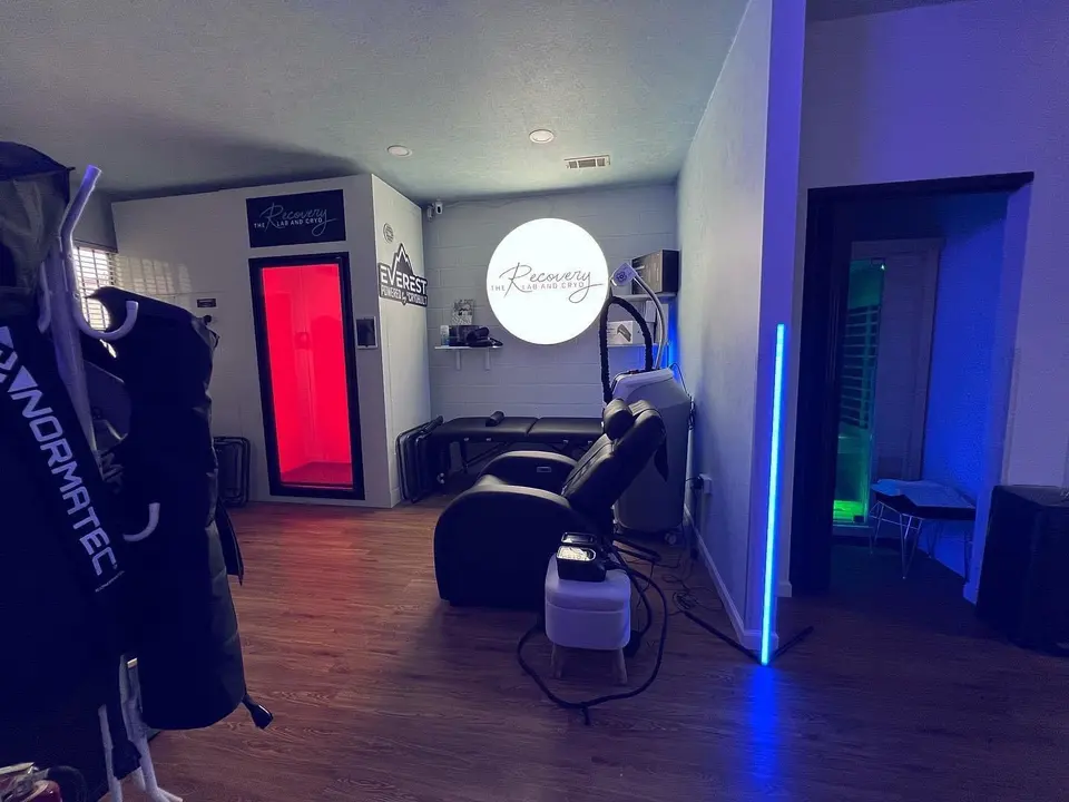 Treatment floor at The Recovery Lab and Cryo with Normatec recovery chairs, a red-lit sauna doorway and the lit-up Recovery logo on the wall